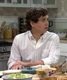 a young man sitting at a kitchen table with food on the counter and bowls in front of him