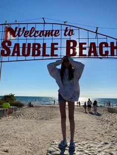 a woman standing under a welcome to sauble beach sign in front of the ocean