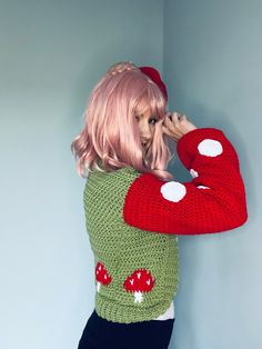 a woman with pink hair wearing a green and red knitted sweater, standing in front of a wall