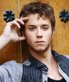 a young man leaning against a wooden door with his hand on his head and looking at the camera