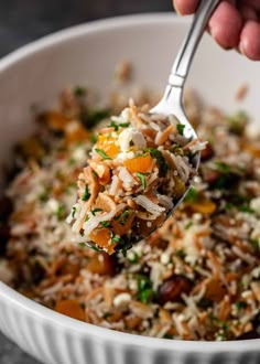 a person holding a spoon full of rice with vegetables and cheese on top, in a white bowl