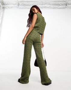 a woman standing in front of a white wall wearing green pants and a black bag