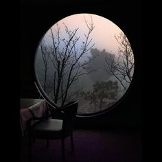 a chair sitting in front of a round window with trees outside the window and fog on the ground