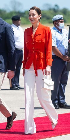 a woman in red jacket and white pants walking down a red carpet next to other people