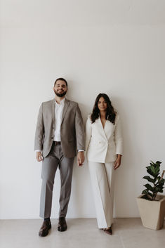 a man and woman standing next to each other in front of a white wall with potted plants