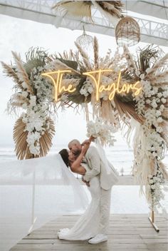 a bride and groom kissing under an arch decorated with white flowers, greenery and feathers