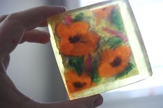 a hand holding up a small square piece of glass with orange flowers painted on it