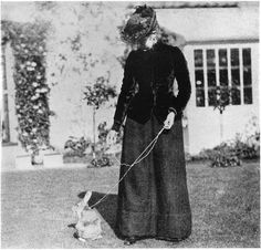 an old black and white photo of a woman with a dog on a leash in front of a house