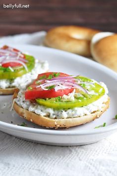 two sandwiches with vegetables and cheese are on a white plate next to bagel buns