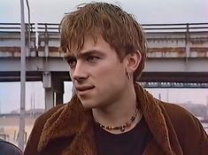 a young man with blonde hair wearing a brown jacket and necklace standing in front of a bridge