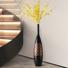 a black vase with yellow flowers in it next to a spiral stair case and wooden blinds