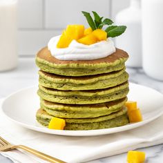 a stack of green pancakes topped with whipped cream and mango slices on a white plate