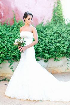 a woman in a white wedding dress standing next to some bushes and plants, holding a bouquet