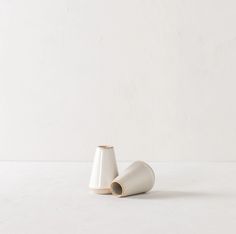 two white vases sitting next to each other on top of a table with a white wall in the background