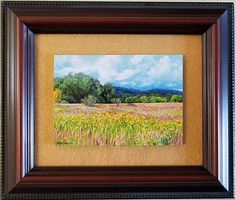 a painting of a field with trees and clouds in the background, framed on a wall