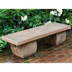 a concrete bench sitting in front of some bushes and flowers on a brick floored walkway