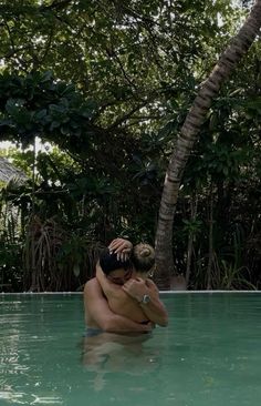 two people are hugging in the middle of a pool surrounded by greenery and trees