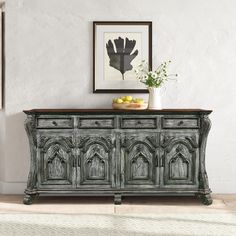 an old dresser with fruit and flowers on top in front of a white wall next to a framed photograph