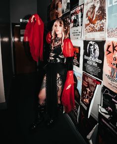 a woman dressed in black and red posing for the camera with posters on the wall behind her