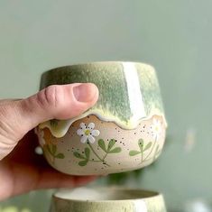 a hand holding a green and white cup with flowers painted on the side, in front of a plant