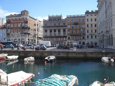 there are many boats that are in the water near some buildings and people walking on the sidewalk