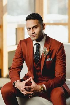 a man in a red suit and tie sitting on a white chair with his hands together