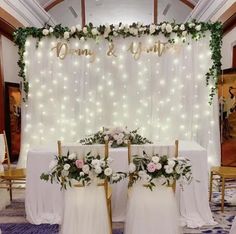 two chairs with flowers and greenery sit in front of a white table cloth covered backdrop