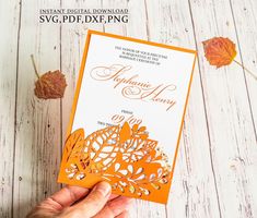 an orange and white wedding card being held up by someone's hand with autumn leaves around it