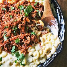 a skillet filled with macaroni and cheese topped with shredded meat next to a wooden spoon