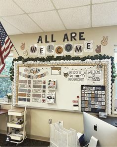 an all are welcome sign on the wall above a computer desk in front of a flag