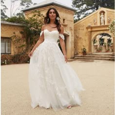 a woman standing in front of a house wearing a wedding dress with an off the shoulder neckline