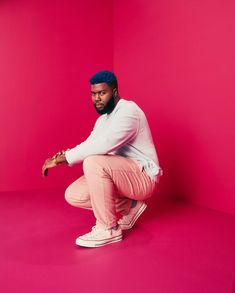 a man squatting on the ground in front of a bright pink background wearing a white sweater and light pink pants