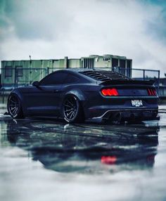 the rear end of a black sports car parked on a wet parking lot with dark clouds in the background