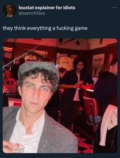 a man with curly hair wearing a gray hat and jacket in a room full of people