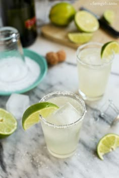 two glasses filled with lemonade and limes on a marble table next to bottles