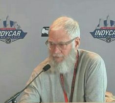 an older man sitting at a table with a microphone in front of him and the indycar logo behind him