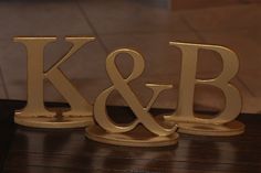 two gold metal letters sitting on top of a wooden table next to each other with the letter k and b in them