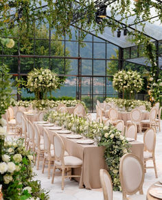 tables and chairs are set up for a formal function in a glass walled area with greenery