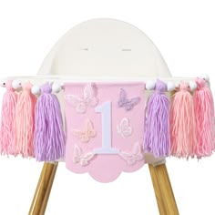 a pink and purple 1st birthday banner with tassels on wooden legs in front of a white background