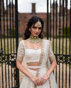 a woman standing in front of a gate wearing a white and gold lehenga