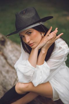 a woman in a white shirt and black hat sitting on a rock with her hands up