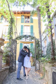 a man and woman are standing in front of a yellow building with green shutters