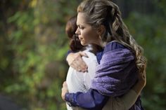 two women hugging each other in front of trees and bushes, one woman is wearing a purple jacket