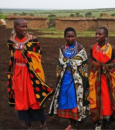 three women in brightly colored clothing standing on dirt ground with grass and trees in the background