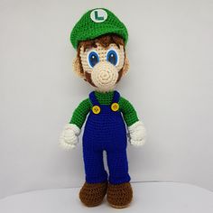 a crocheted stuffed toy with a green hat and blue overalls is posed on a white surface