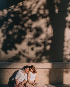 a man and woman sitting on the ground next to each other under a tree in front of a building