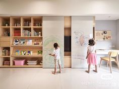 two young children are drawing on the wall in their playroom, with bookshelves behind them