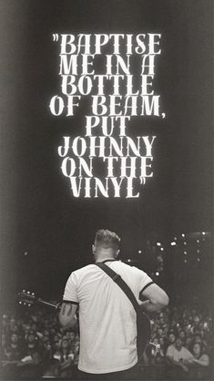 a man holding a guitar in front of a large screen with words written on it