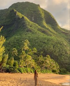 a woman standing on top of a sandy beach next to a lush green mountain covered in trees