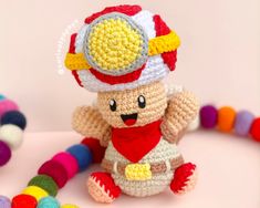 a crocheted teddy bear wearing a hat and holding a bead necklace on a pink surface